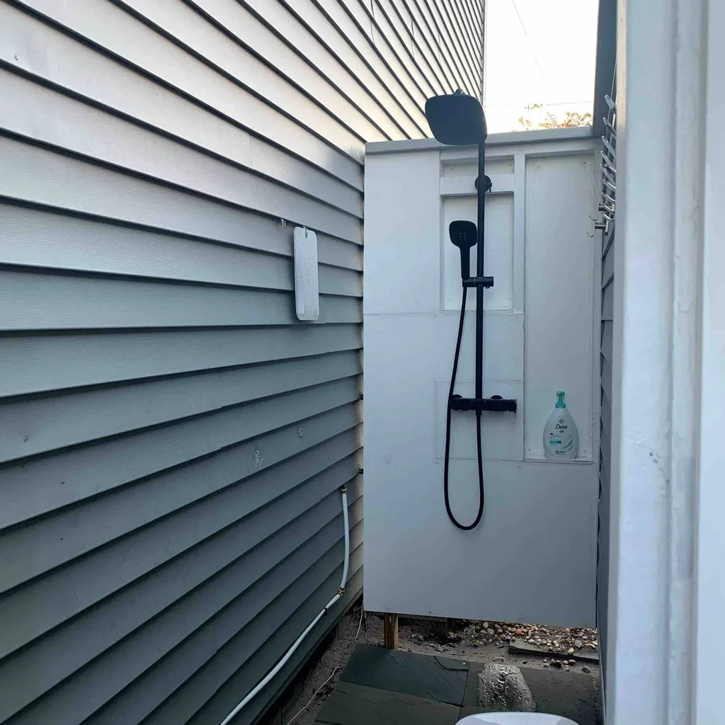 Outdoor shower beside gray siding, with black overhead and handheld showerheads mounted on a white panel.