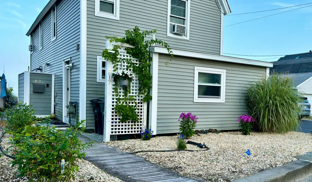 Gray-sided house with white trim, a vine-covered lattice, flowering plants and a wooden walkway.
