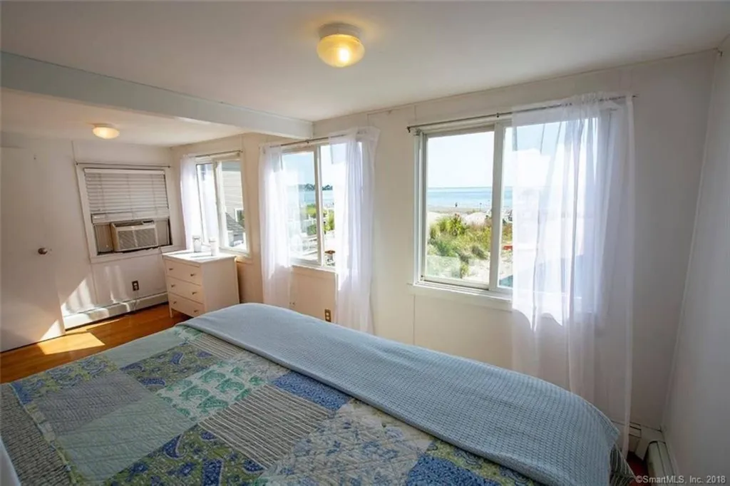 Bedroom with a blue patchwork quilt, sheer white curtains, window AC and views toward the water.